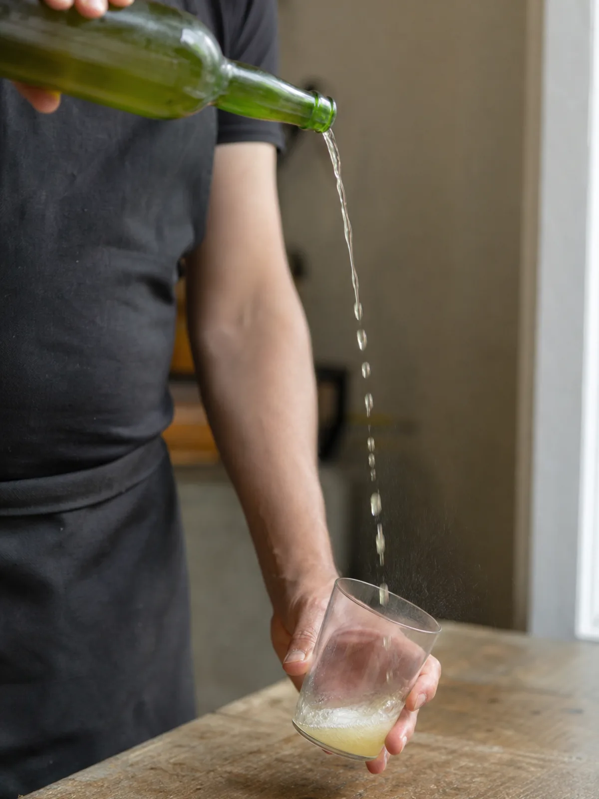 A person pouring cider from a bottle held overhead into a wide glass held low at their side.